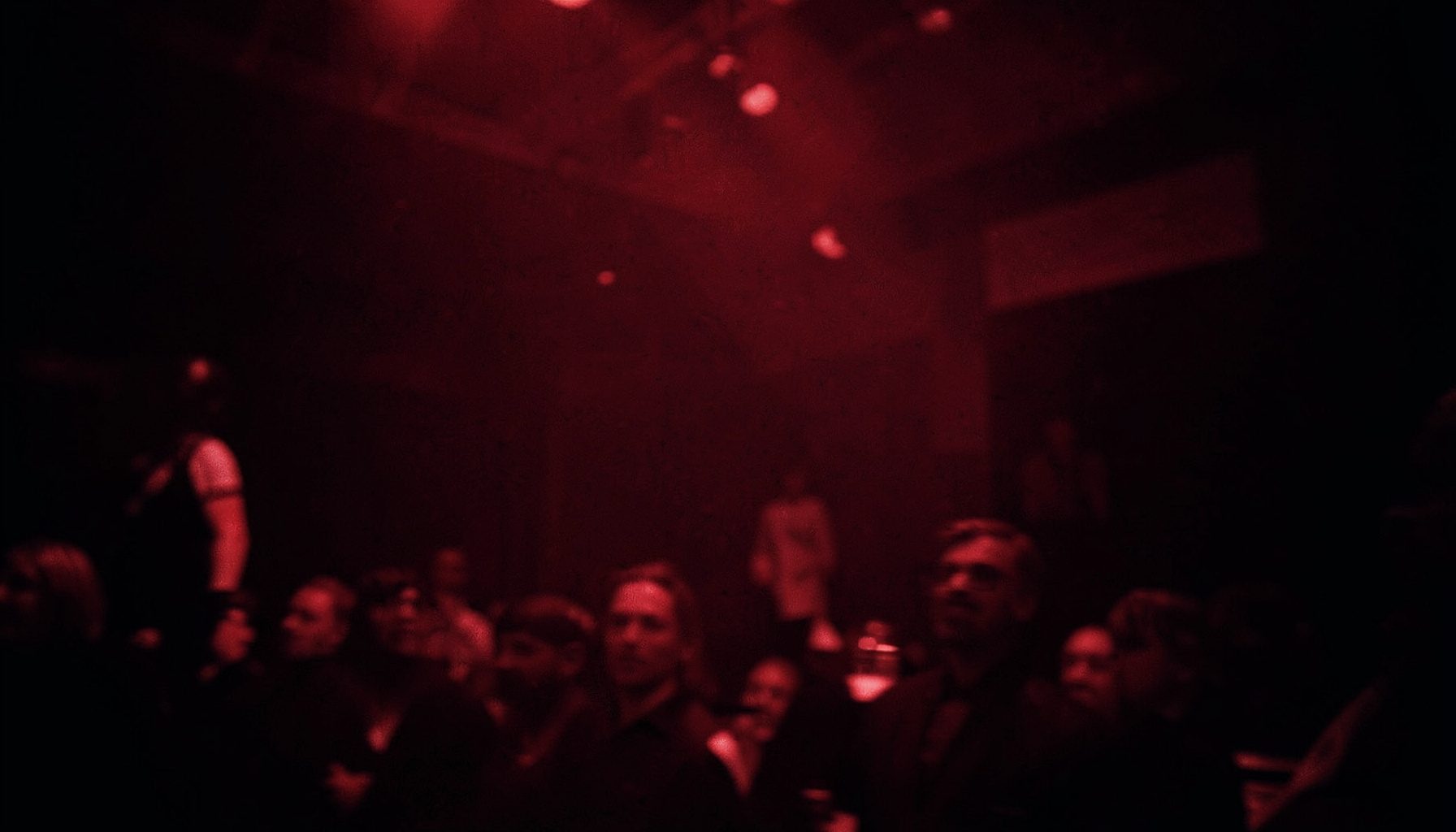 Dark atmospheric duotone photograph with deep red and black tones, suggesting a dimly lit performance space with subtle motion blur and dramatic shadows