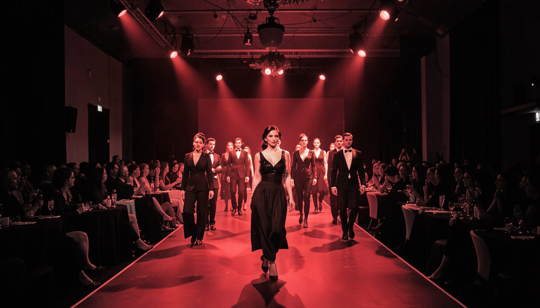 Duotone red-and-black photograph of a ballroom scene with contestants walking the runway under spotlights