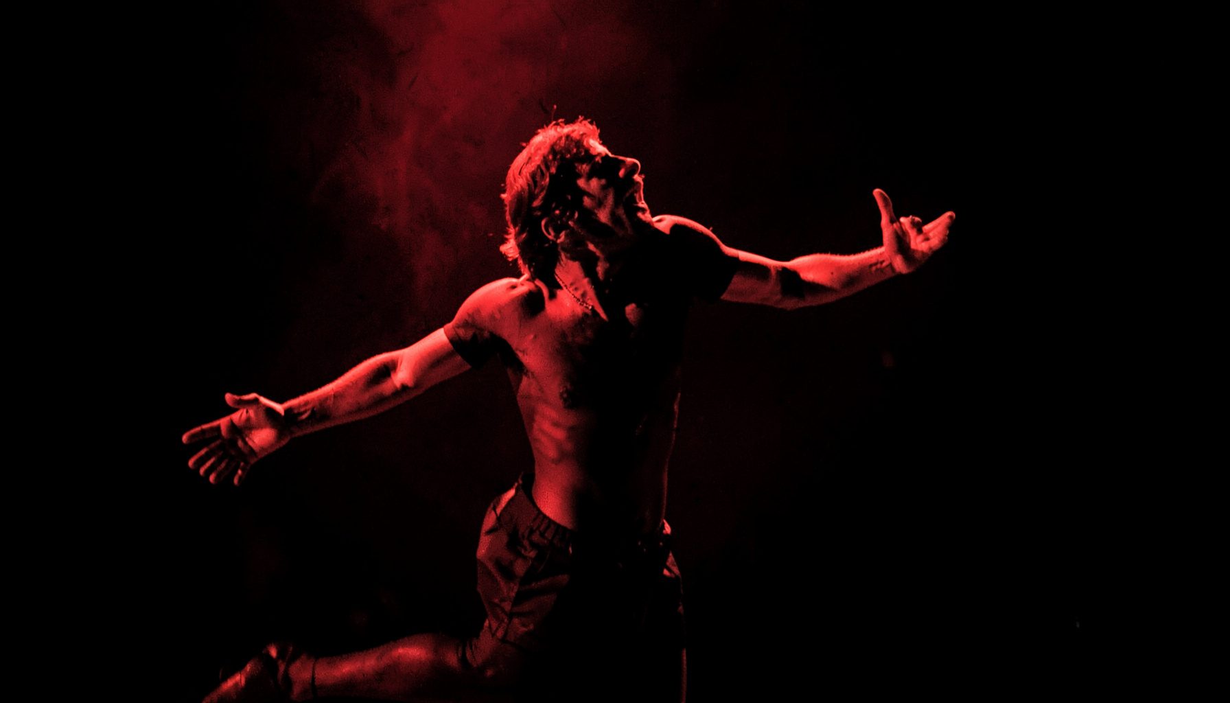 Duotone photograph in red and black showing a performer mid-motion on a dim stage, arms extended with dramatic lighting