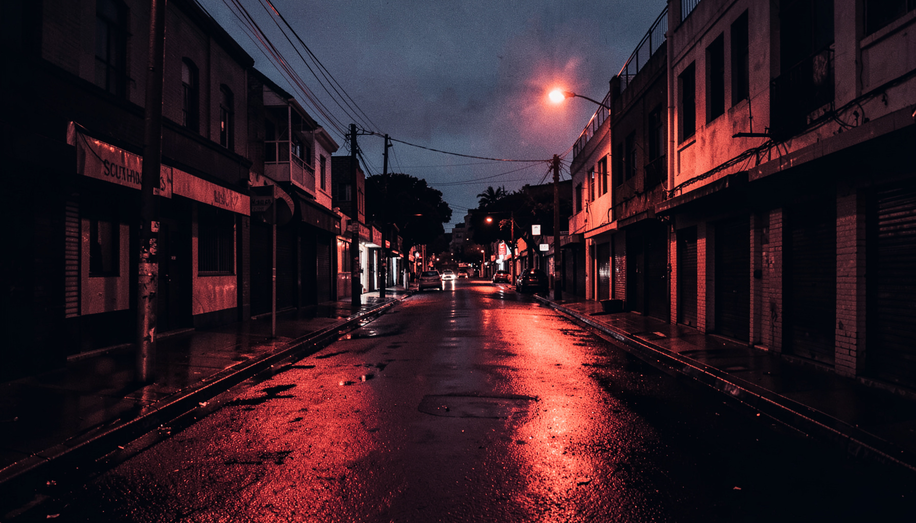 A dark duotone photograph of an urban South Auckland street at dusk, warm amber streetlights reflecting off wet pavement, deep navy shadows
